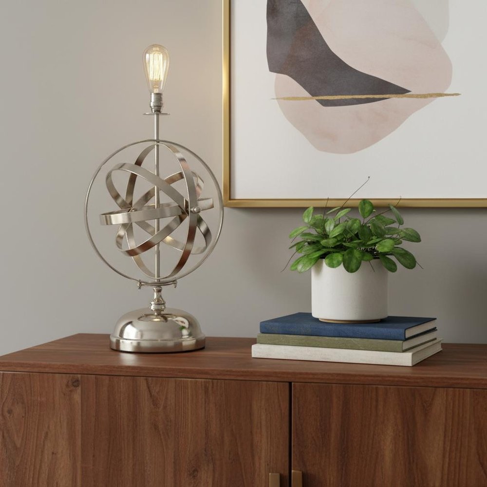 Bronze metal standard table lamp with exposed bulb on a wood sideboard, white planter with greenery atop books, abstract gold-framed art.