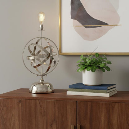 Bronze metal standard table lamp with exposed bulb on a wood sideboard, white planter with greenery atop books, abstract gold-framed art.