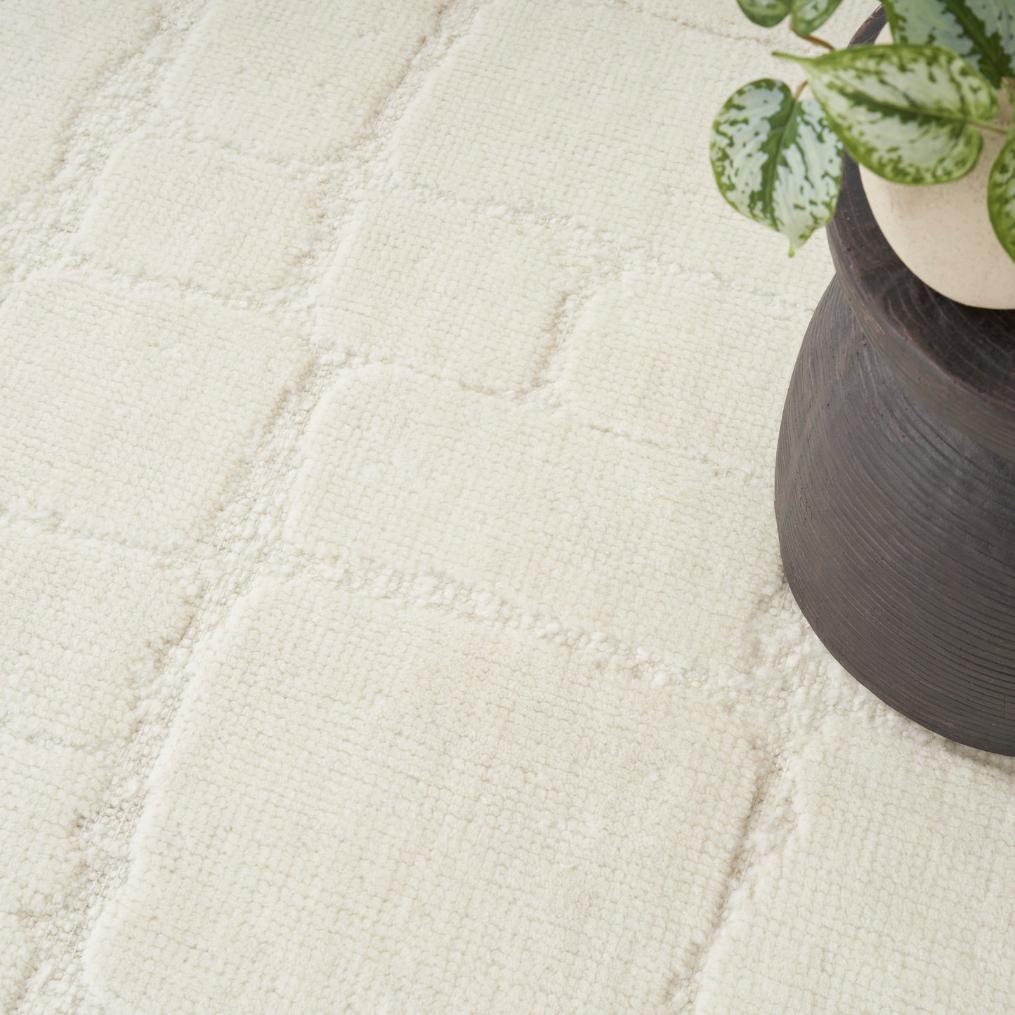 Cream textured rug with square tufted blocks; neutral decor beside a dark planter with a green variegated plant.