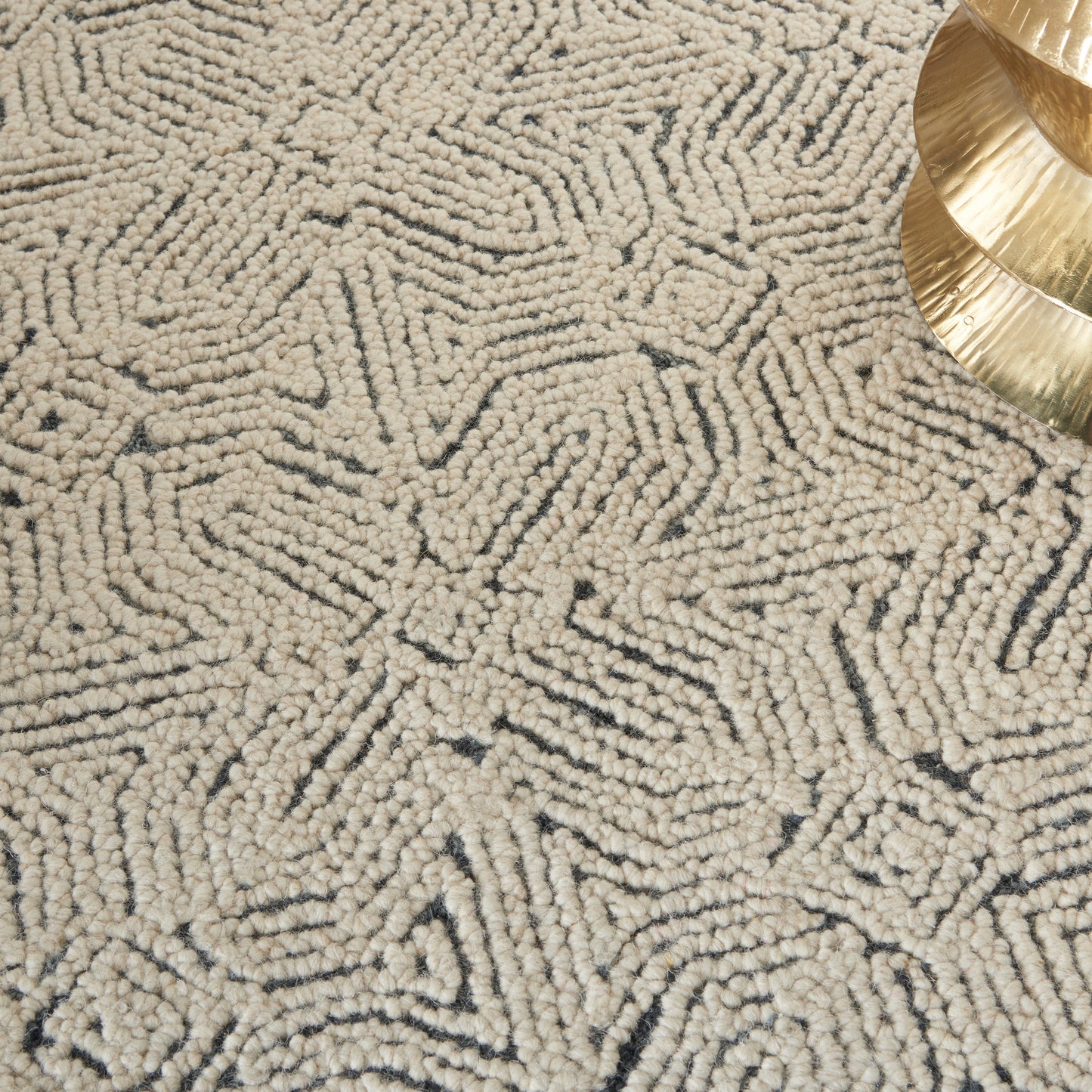 Beige loop-pile area rug with a dark geometric line pattern and a gold decorative piece in the corner.