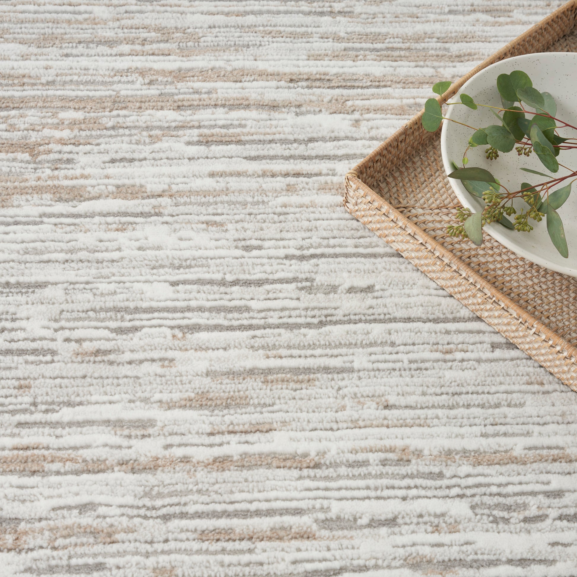 Beige and cream striped textured rug with a woven tray, white bowl, and eucalyptus.