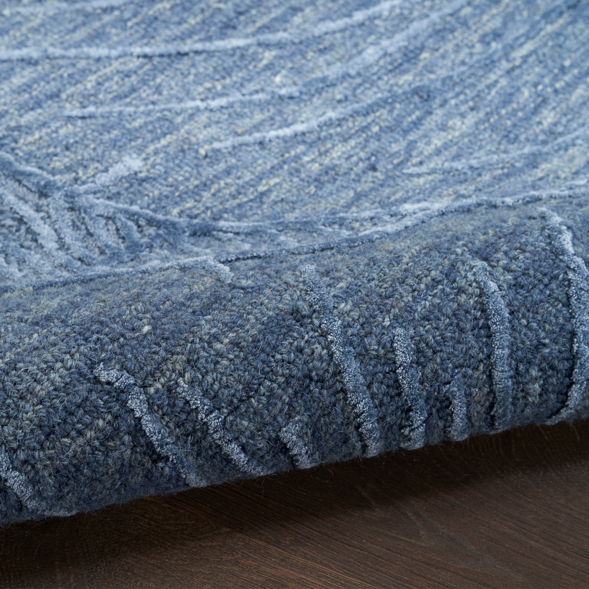 Blue textured area rug with a chunky loop pile and abstract lighter-blue pattern, rolled edge on a wooden floor.