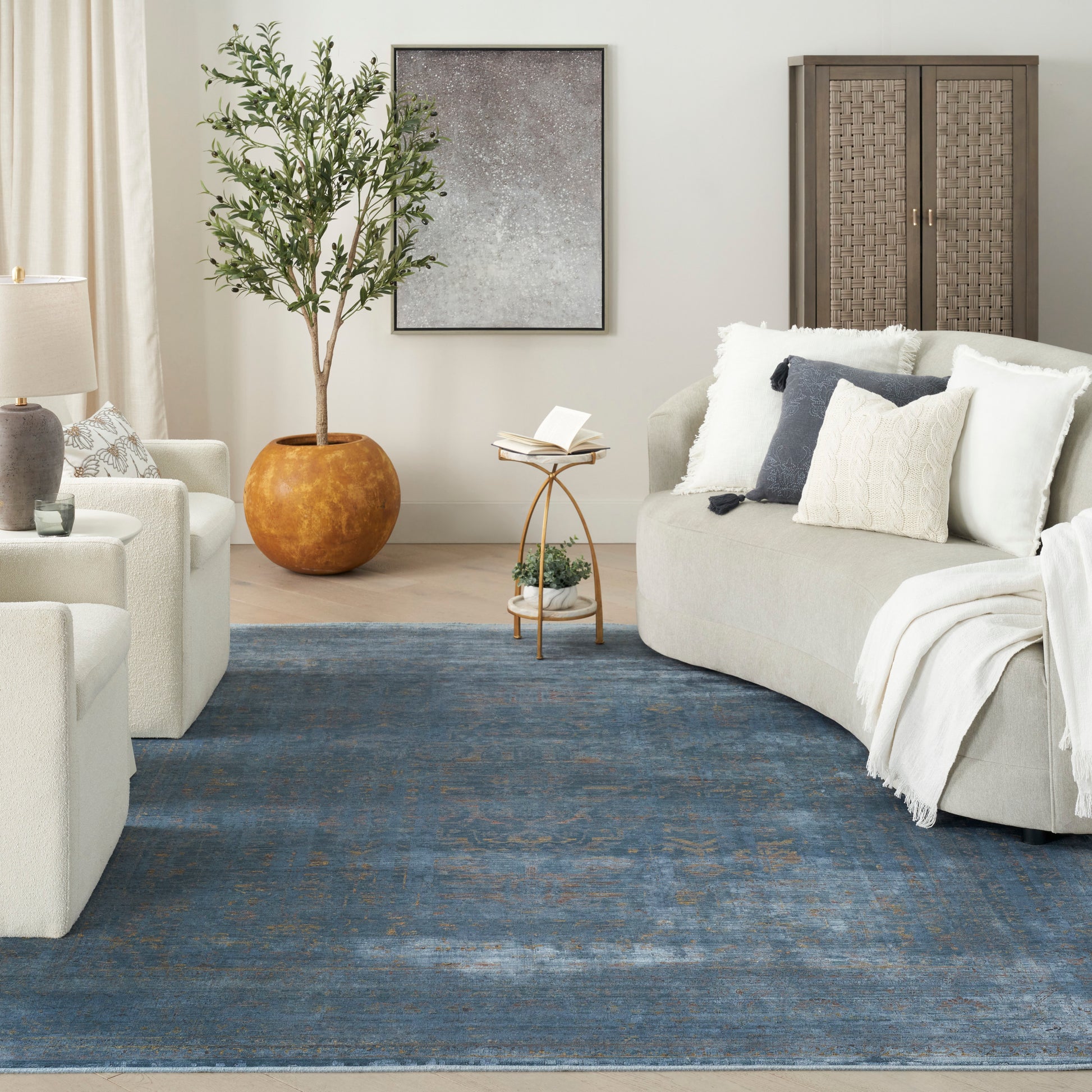 Cozy living room with a blue rug, cream sofas, neutral pillows, olive tree in a terracotta pot, and woven cabinet.