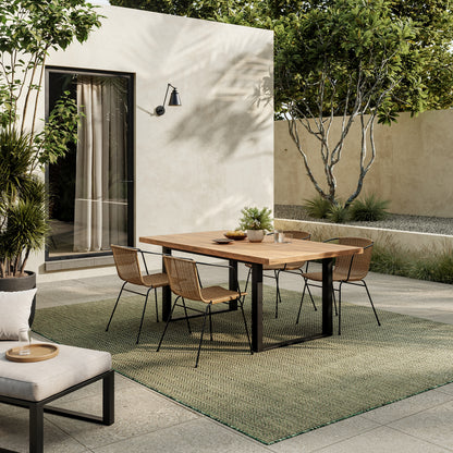Modern outdoor dining set: rectangular wooden table with black metal legs, six wicker chairs on a green rug beside a beige wall and plants.