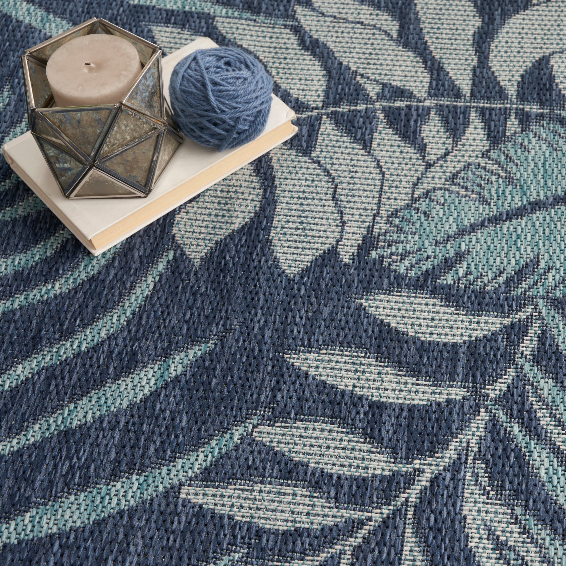 Geometric glass candle holder with beige candle on a book, blue yarn nearby on a blue-teal patterned rug.