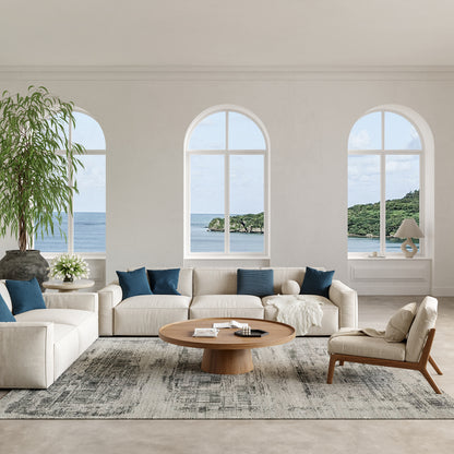 Bright coastal living room with a modern 10' X 14' rug, beige fabric sofas & blue cushions, round wooden coffee table, light armchair & large arched windows overlooking the sea.