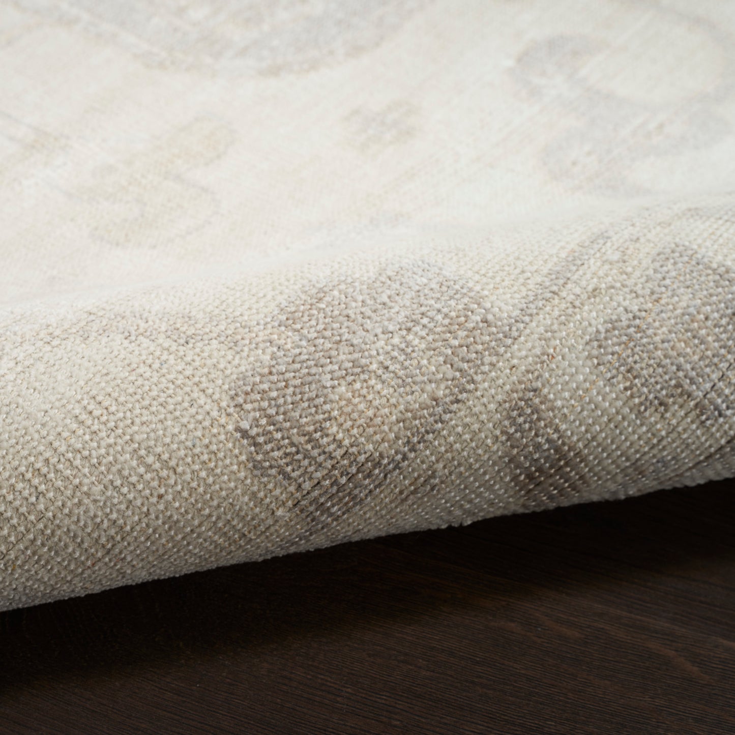 Beige woven rug close-up with subtle damask floral pattern; textured fabric folded over dark wooden floor.