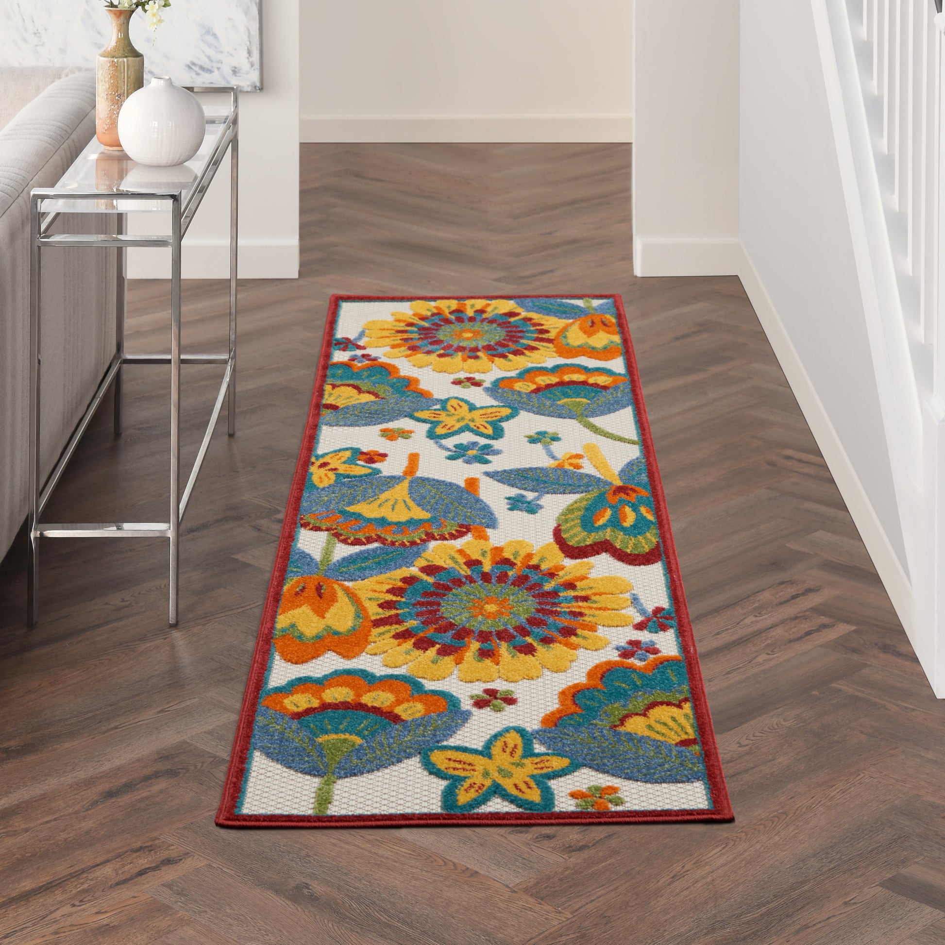 Long colorful floral runner rug with red border on herringbone wood floor in hallway beside glass console table.
