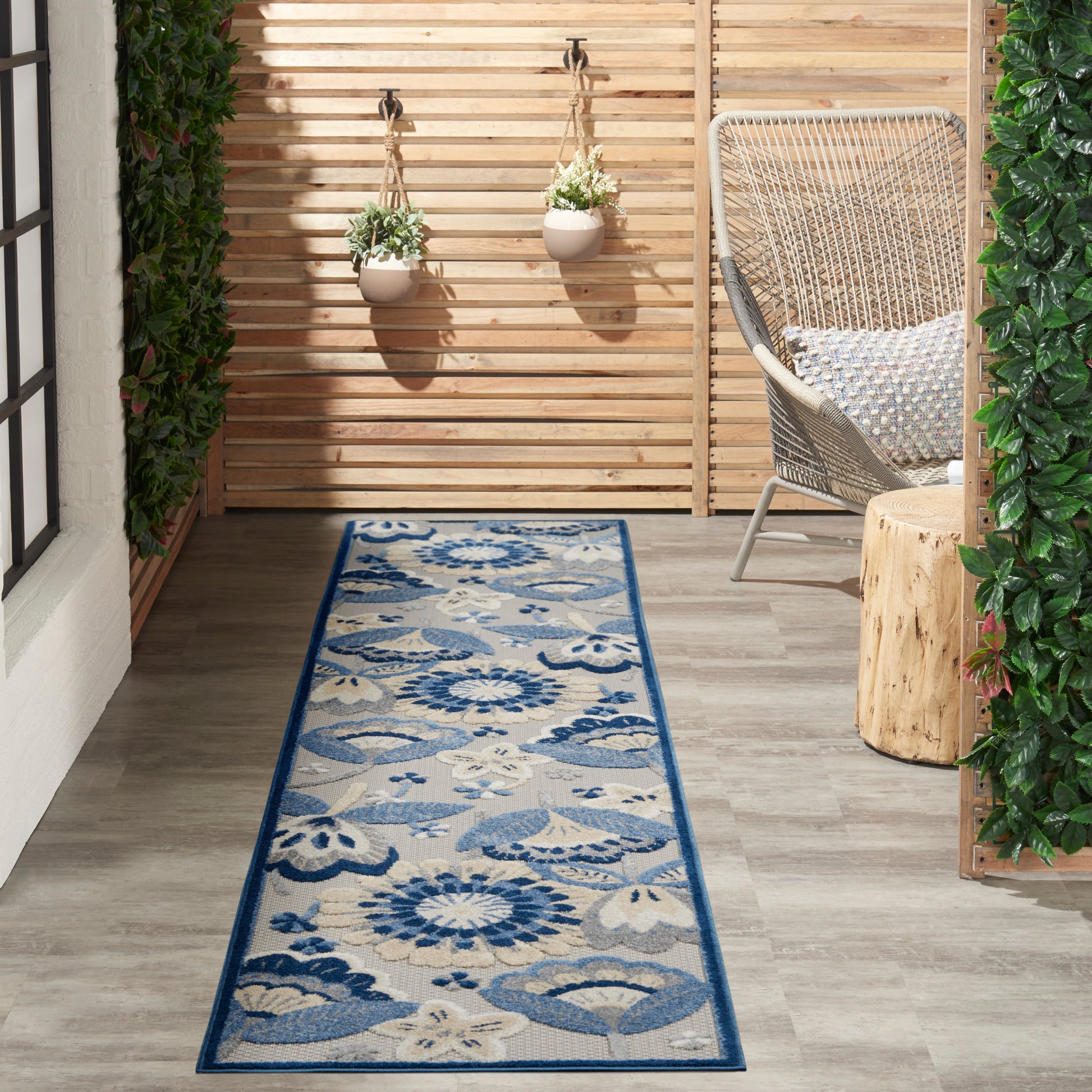 Outdoor patio with wooden slat wall, blue floral runner rug, woven wicker chair, hanging planters, and greenery.