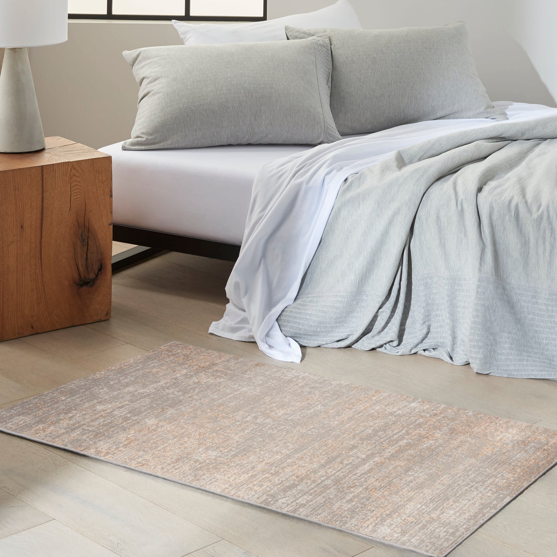 Soft gray bedding with white sheets and two gray pillows; wooden nightstand and beige-toned runner rug on pale wood floor.