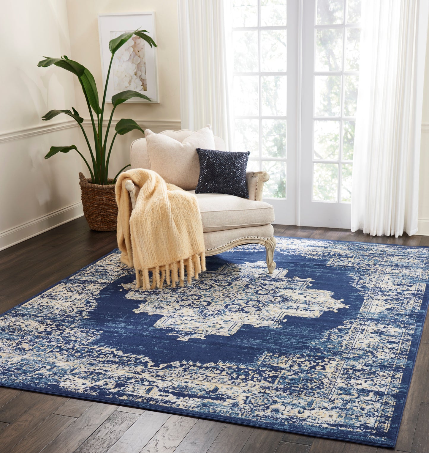 Cream tufted armchair with beige chunky throw and navy sequin pillow on a distressed blue-and-ivory rug; plant by window.