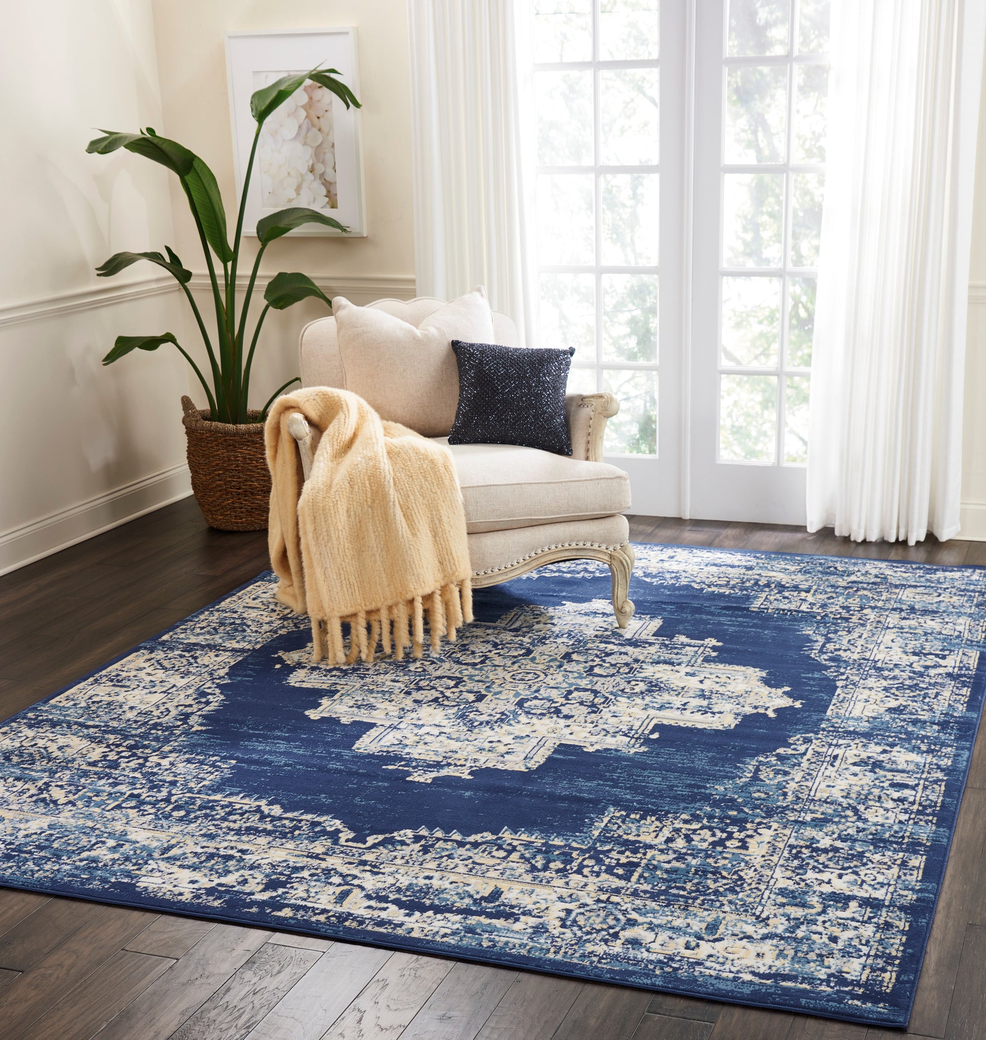 Cream tufted armchair with beige chunky throw and navy sequin pillow on a distressed blue-and-ivory rug; plant by window.