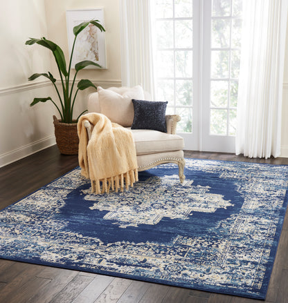 Cream tufted armchair with beige chunky throw and navy sequin pillow on a distressed blue-and-ivory rug; plant by window.