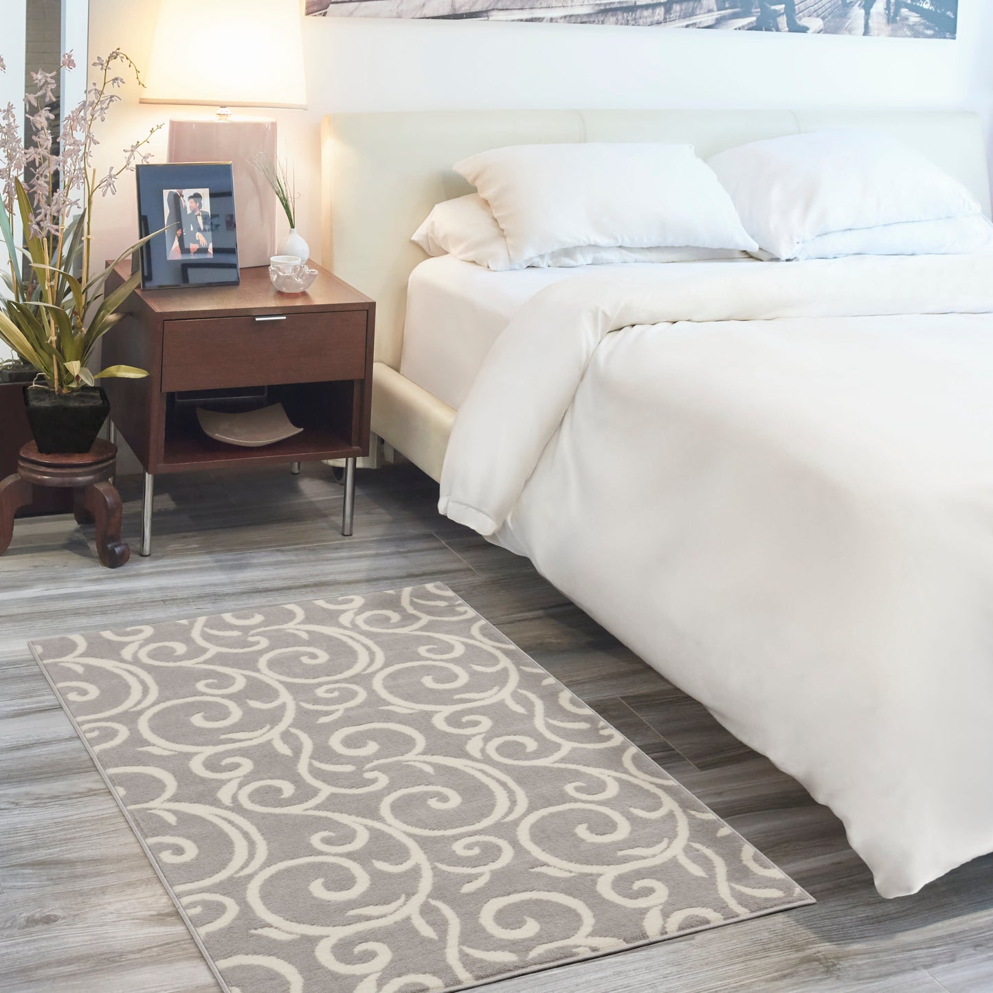 White bed with plush comforter & pillows, dark wood nightstand, lamp, framed photo, plant; gray swirl rug on wood floor.