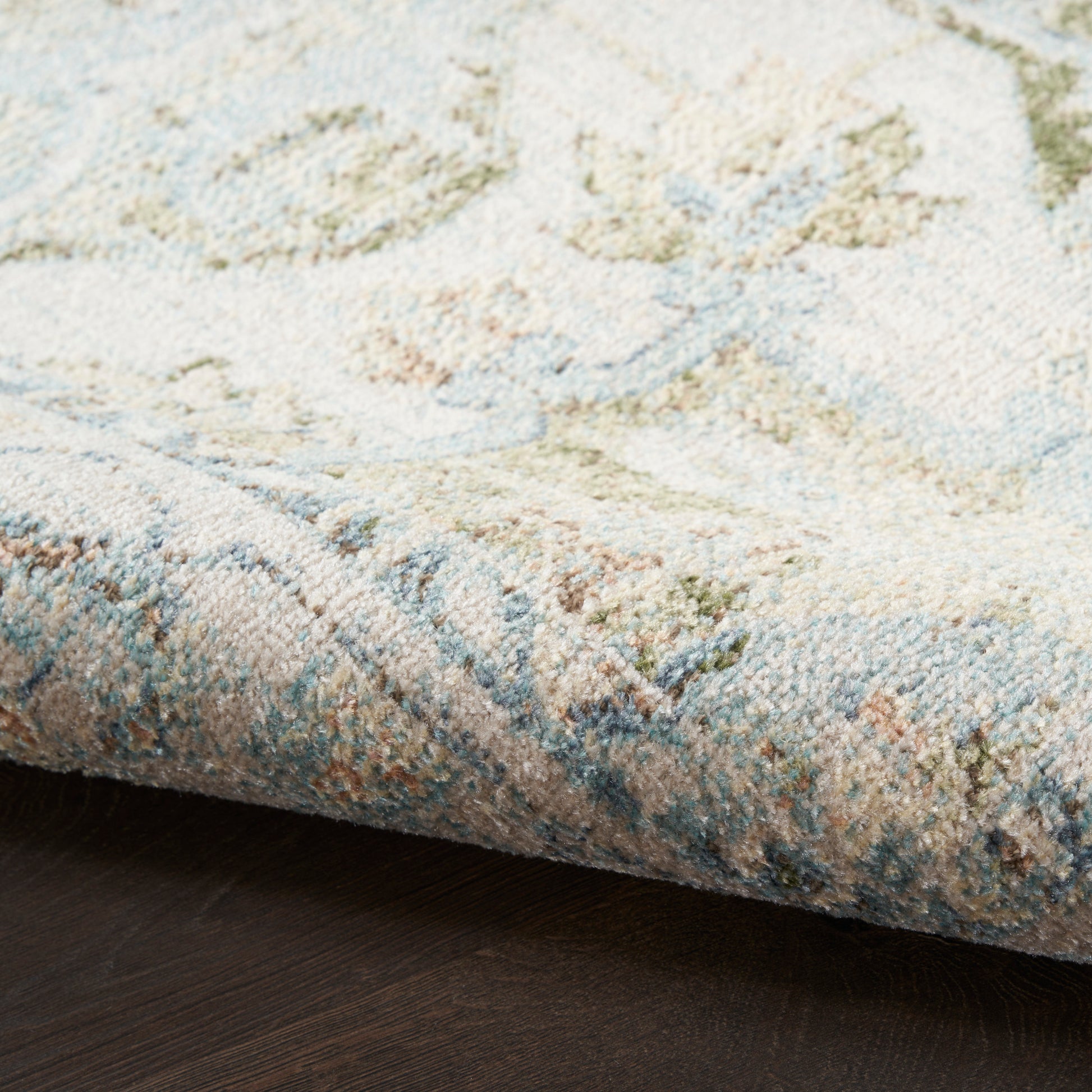 Close-up of plush pastel rug with blue, beige and sage green tones; folded edge on dark wood floor.