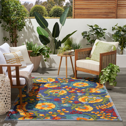 Outdoor patio scene with a vibrant blue floral rug, wooden-frame chairs with cream cushions, and lush potted plants.