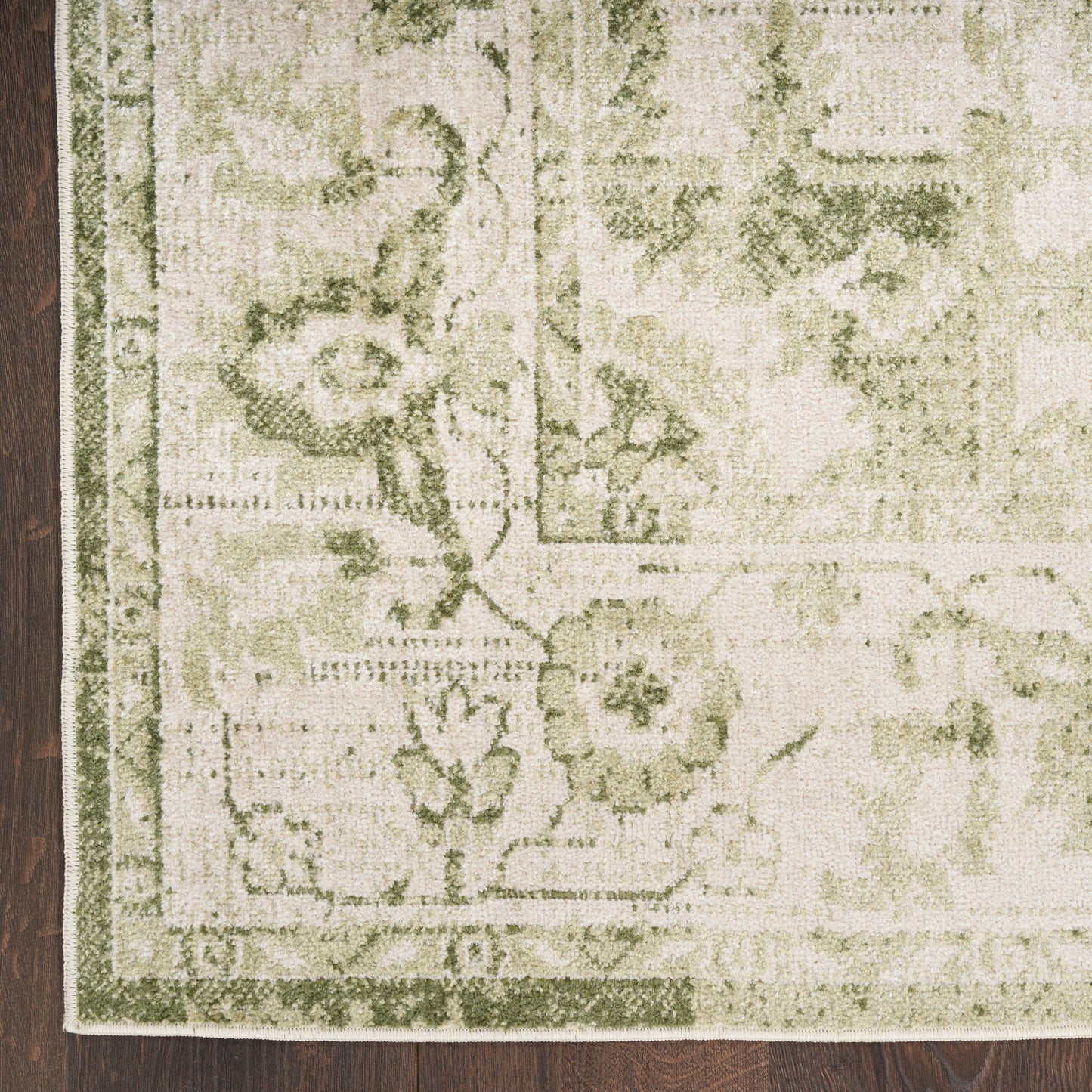 Beige rug with sage green floral pattern and border on dark wood floor; fringe edge visible.