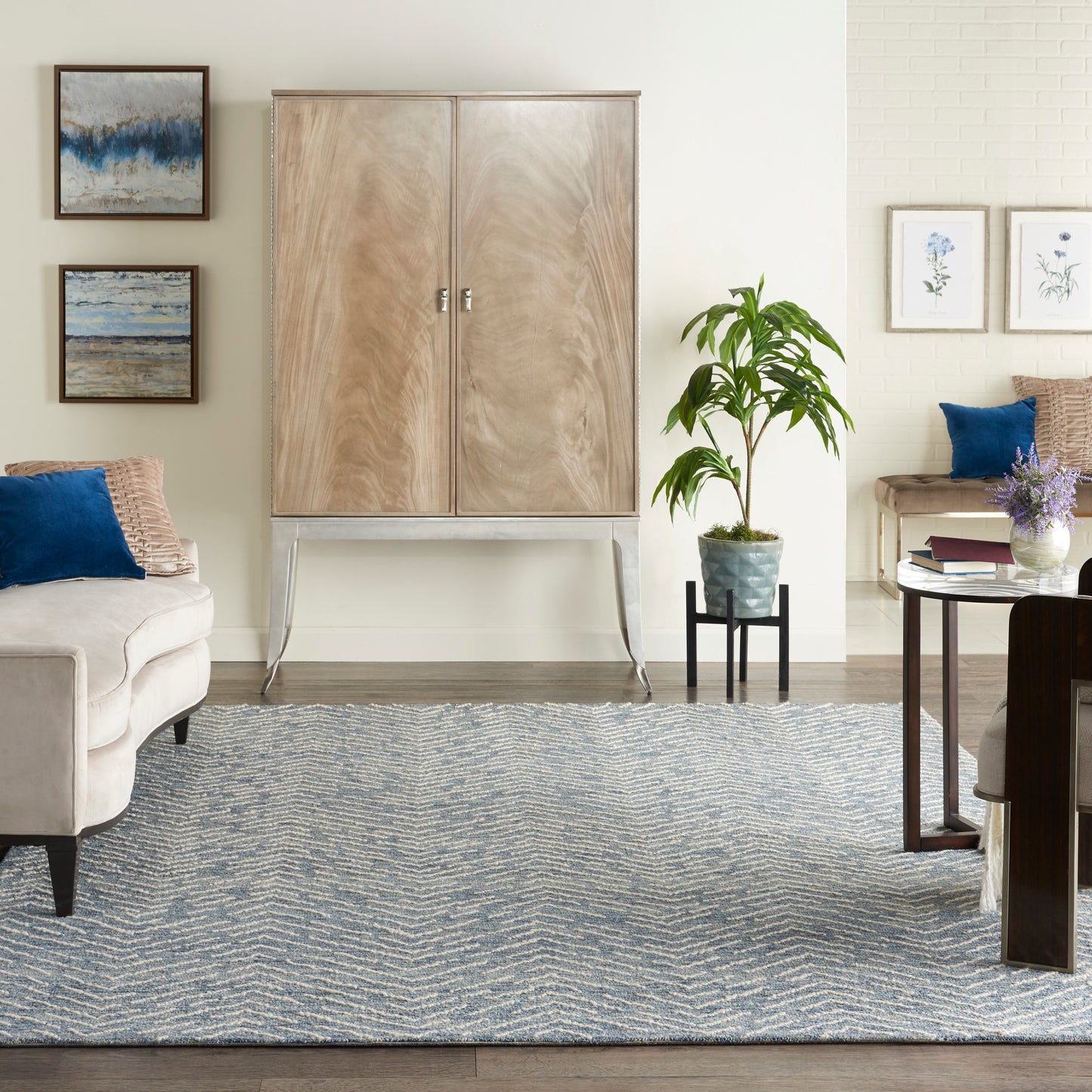 Light wood armoire on metal legs in a neutral living room with blue pillows, zigzag rug, plant, and botanical art.