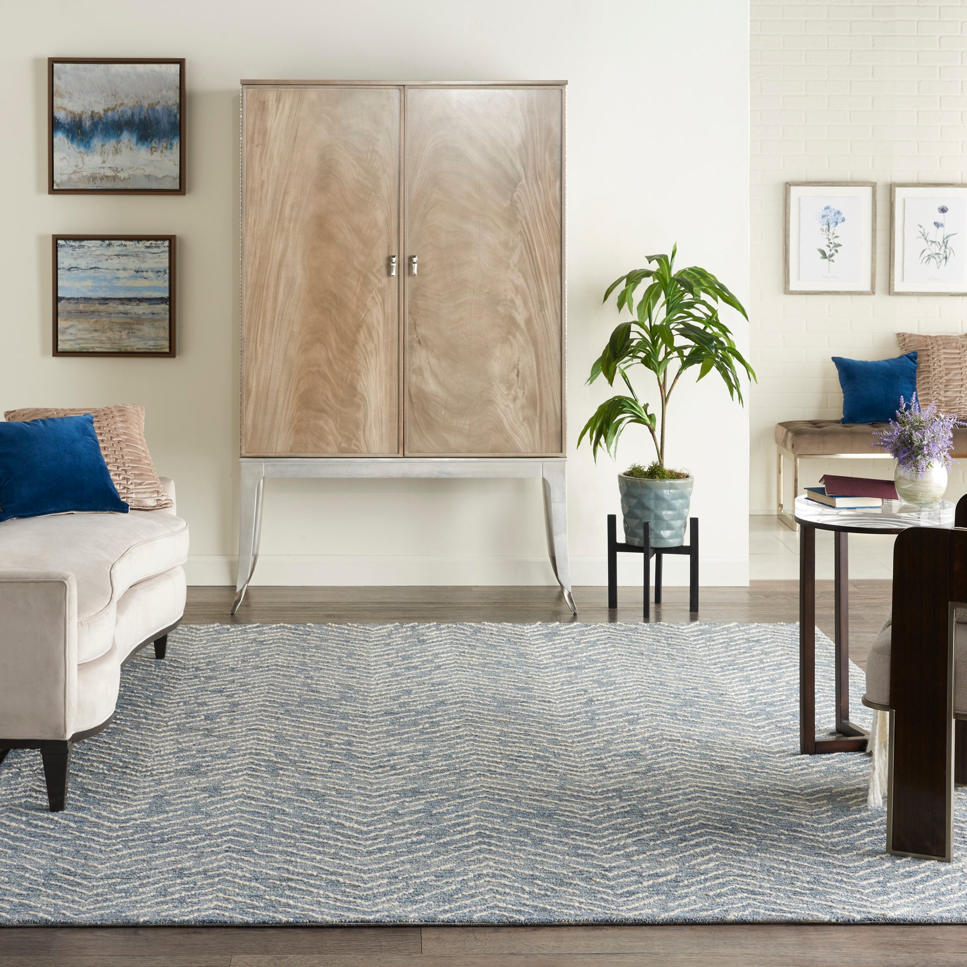 Light wood armoire on metal legs in a neutral living room with blue pillows, zigzag rug, plant, and botanical art.