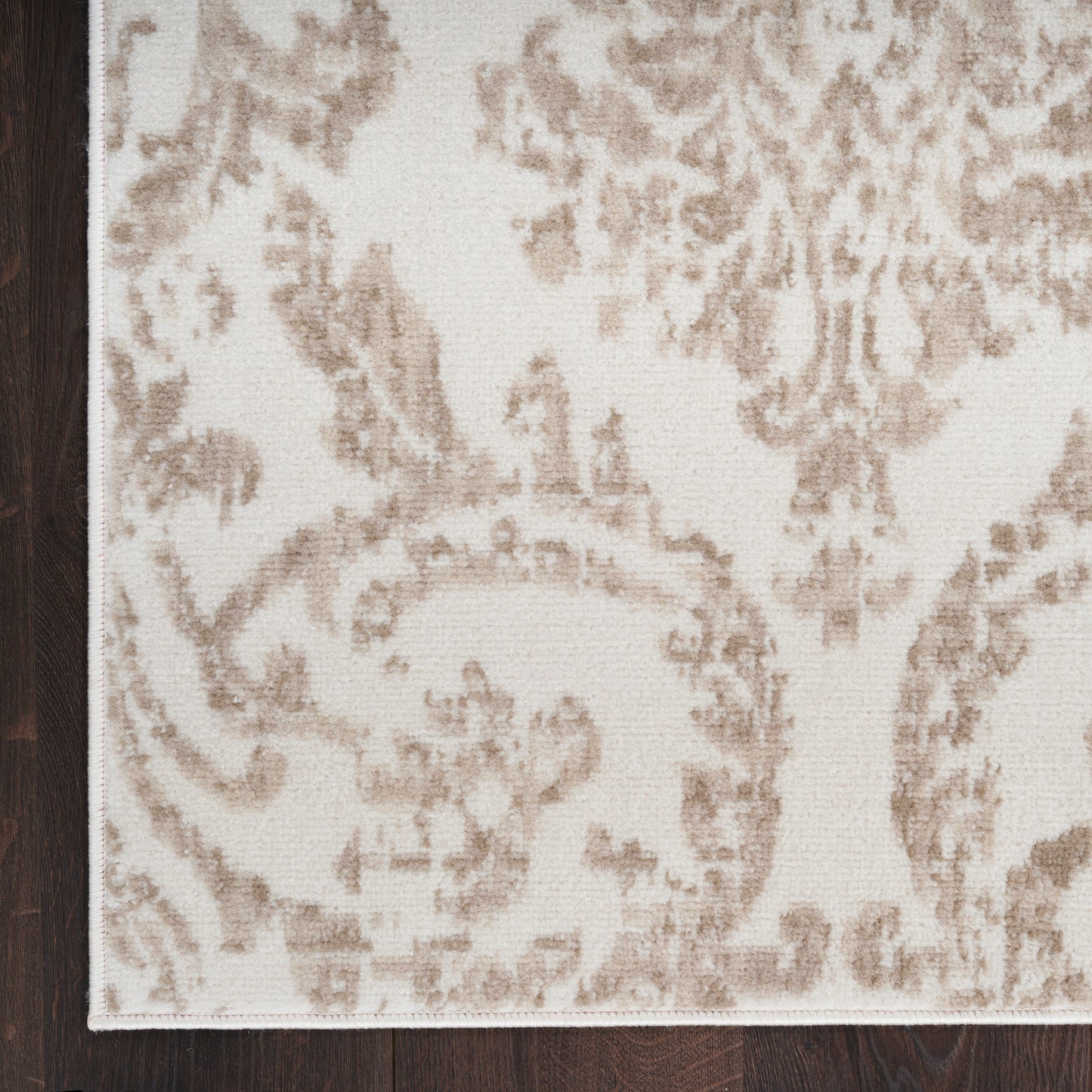 Corner view of a beige damask-pattern rug with light brown floral scrolls on a dark wood floor.