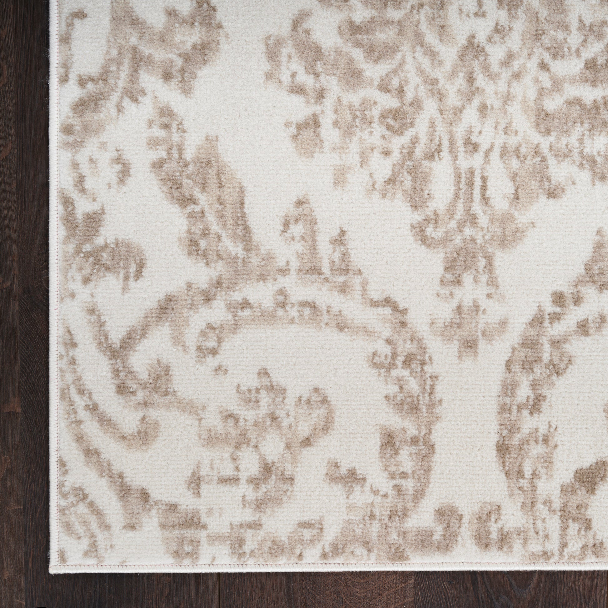 Corner view of a beige damask-pattern rug with light brown floral scrolls on a dark wood floor.