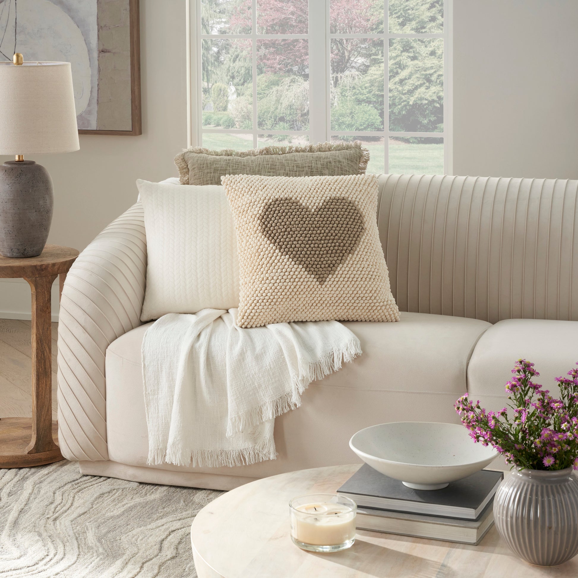 Cream striped sofa with heart pillow, beige cushion, cream throw; wood side table, lamp, round coffee table with bowl.