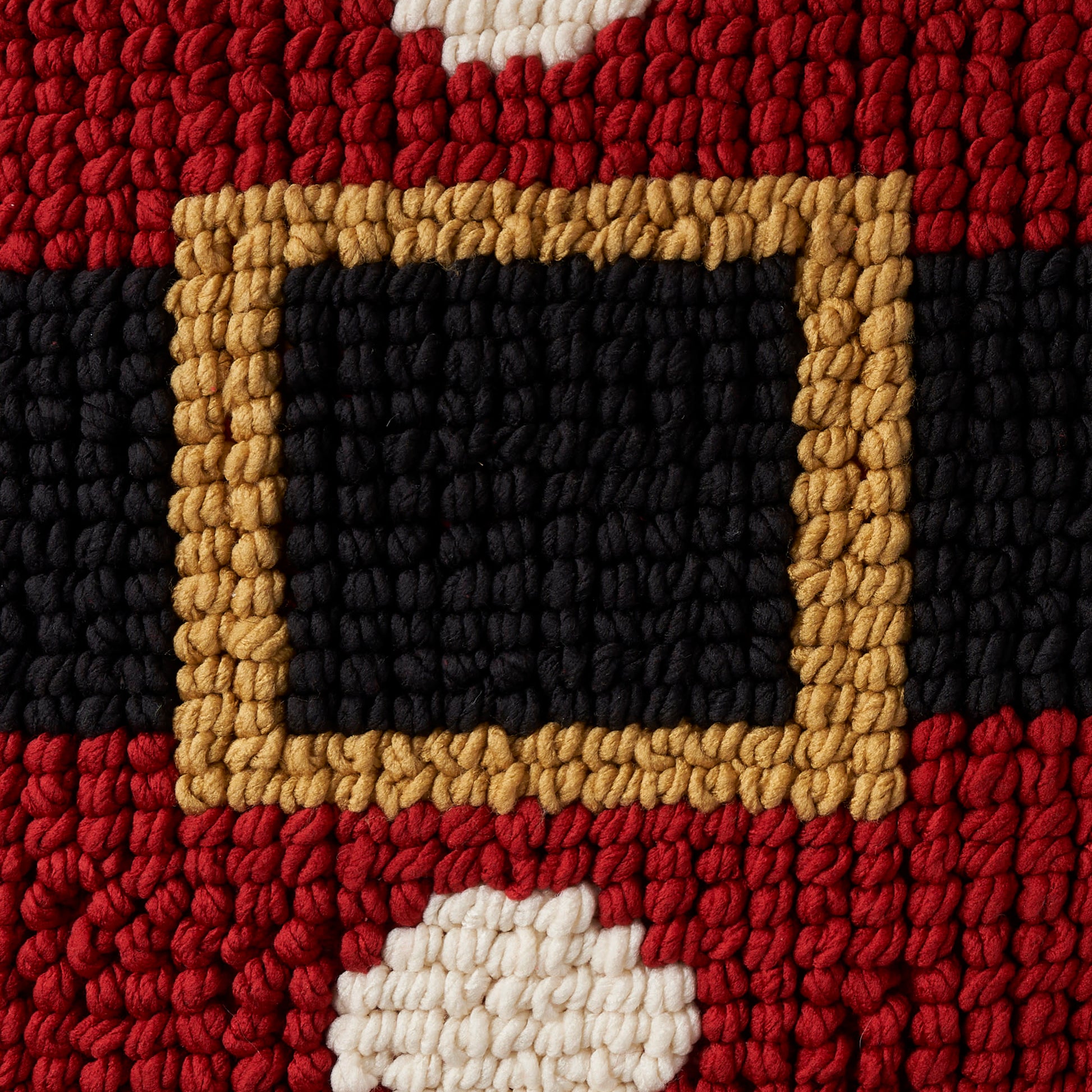 Chunky woven rug close-up with red and black bands and a tan belt-buckle frame around a black center, white tuft accents.