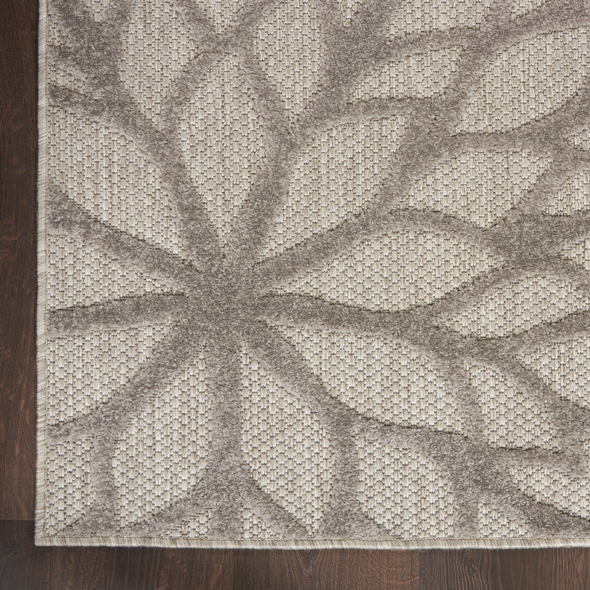 Beige and taupe textured rug with a large leaf/starburst pattern and a fringed edge on dark wood floor.