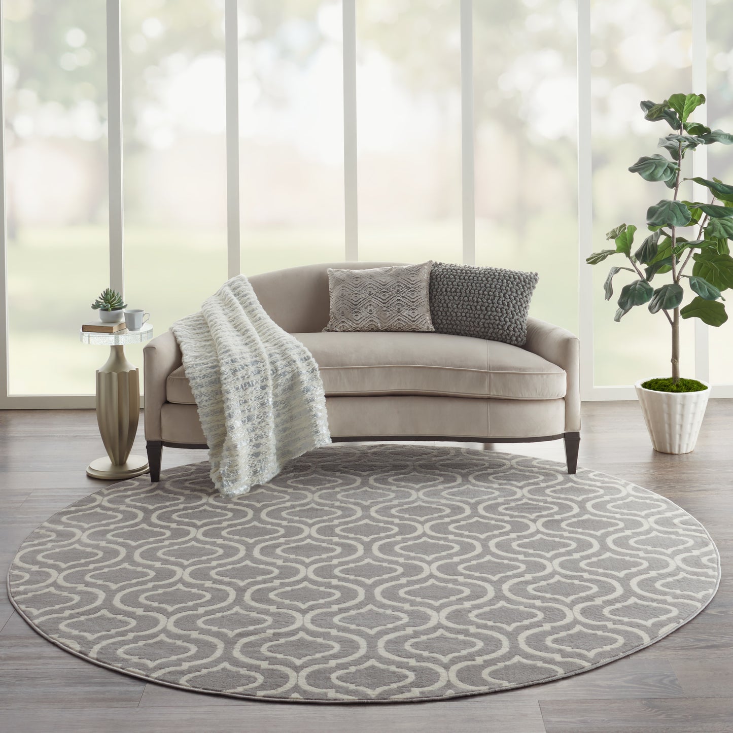 Round gray rug with white swirl under beige sofa; knitted throw, two pillows, gold side table, potted plant by window.