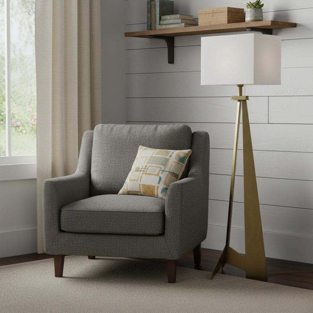 Gray fabric armchair with a patterned beige-yellow pillow, beside a gold tripod floor lamp in a shiplap-walled living nook.