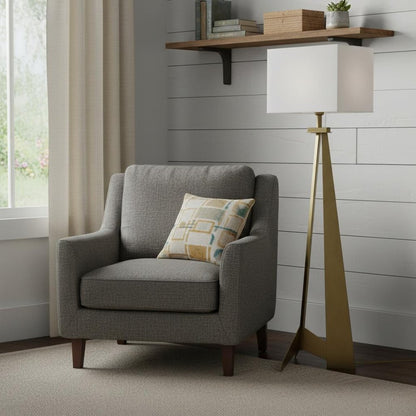 Gray fabric armchair with a patterned beige-yellow pillow, beside a gold tripod floor lamp in a shiplap-walled living nook.