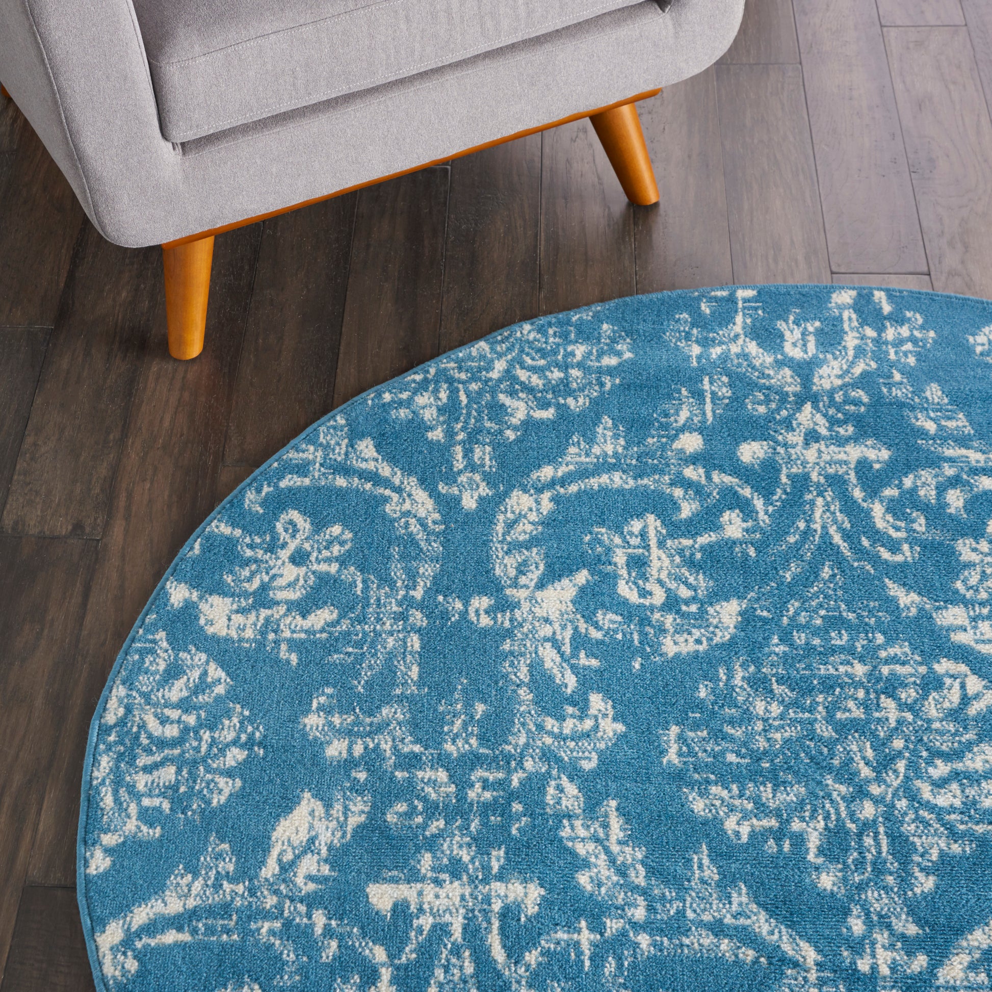 Round blue rug with a white distressed pattern, under a gray sofa with orange legs on dark wood flooring.