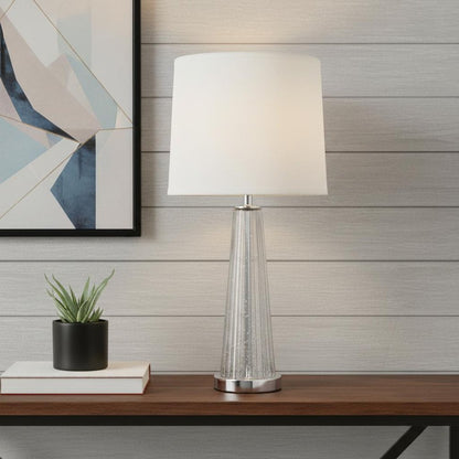 Clear glass tapered pedestal lamp with an Empire shade on a wood console, beside a succulent in a black pot and abstract art.