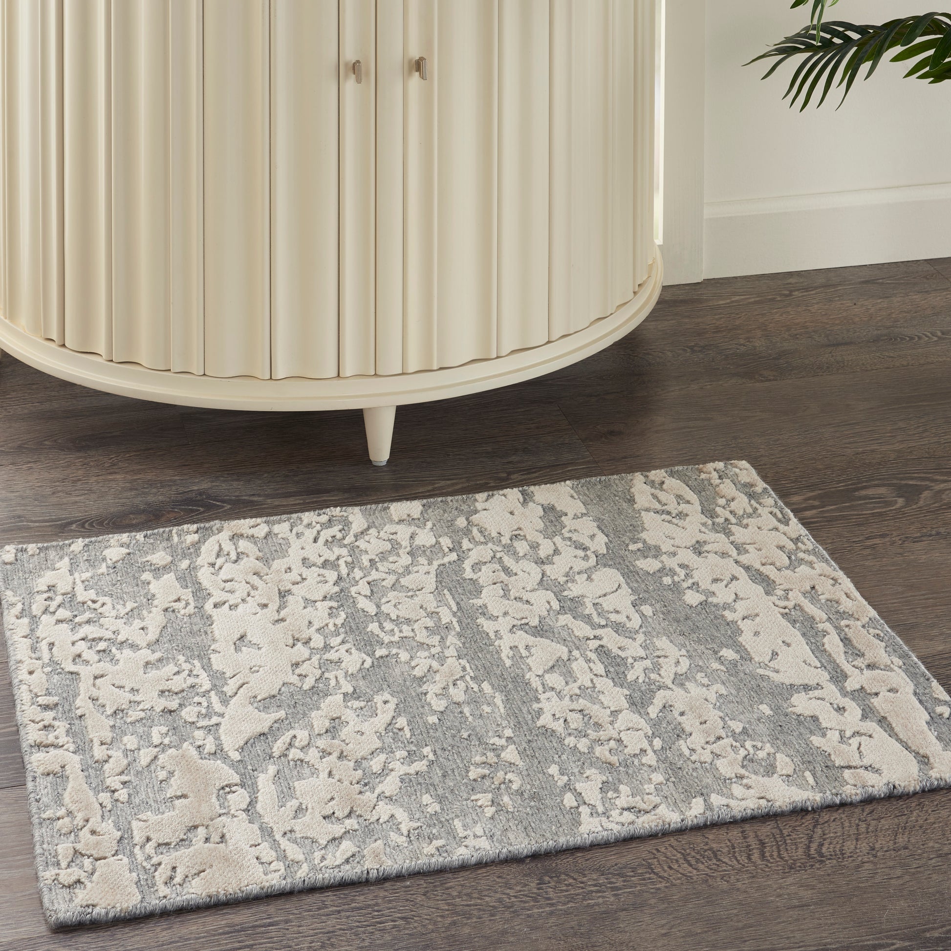 Beige curved ribbed cabinet on dark wood floor beside a gray patterned rug; plant peeks in.