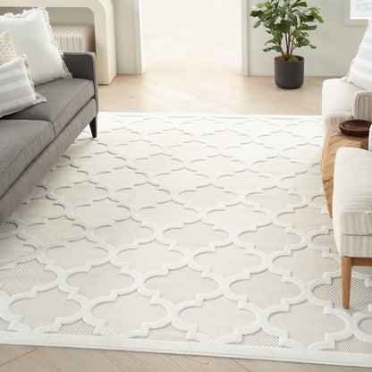 Cream geometric trellis rug in a bright living room with gray sofa, white cushions, light wood floors, & a potted plant.