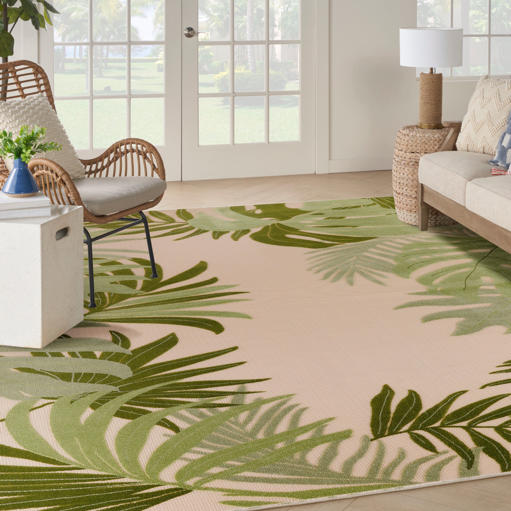 Beige outdoor rug with a vivid green tropical palm leaf pattern, in a sunlit space with a wicker chair and rattan side table.