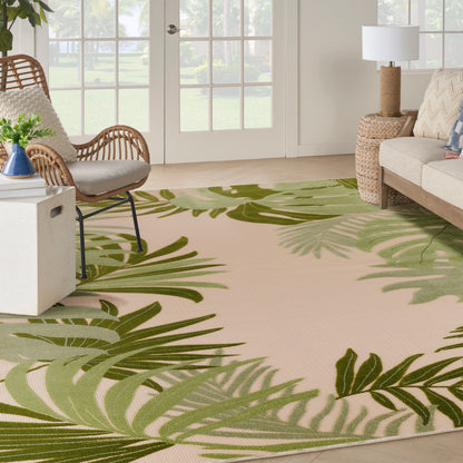 Beige outdoor rug with a vivid green tropical palm leaf pattern, in a sunlit space with a wicker chair and rattan side table.