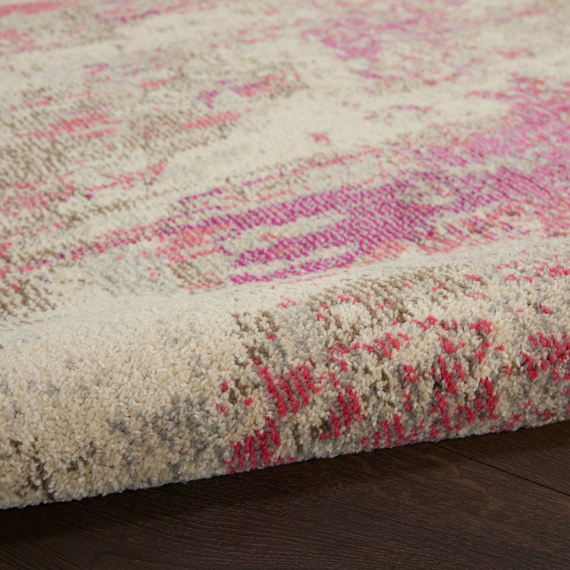 Beige plush area rug with pink abstract pattern; tufted pile and visible edge on dark wood floor, contemporary style.
