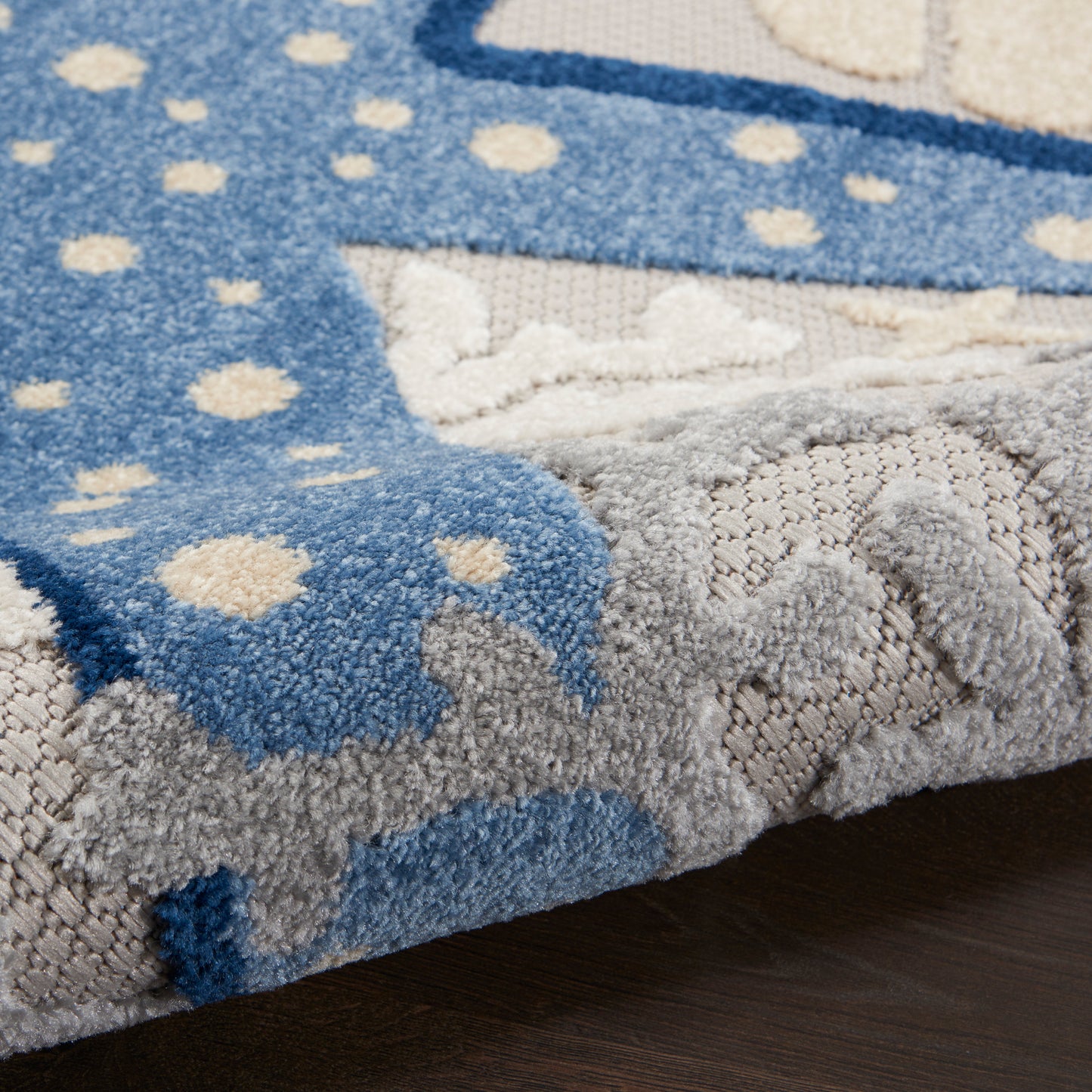 Close-up of blue tufted area rug with cream dots, folded edge revealing soft pile and gray underside on dark wood floor.