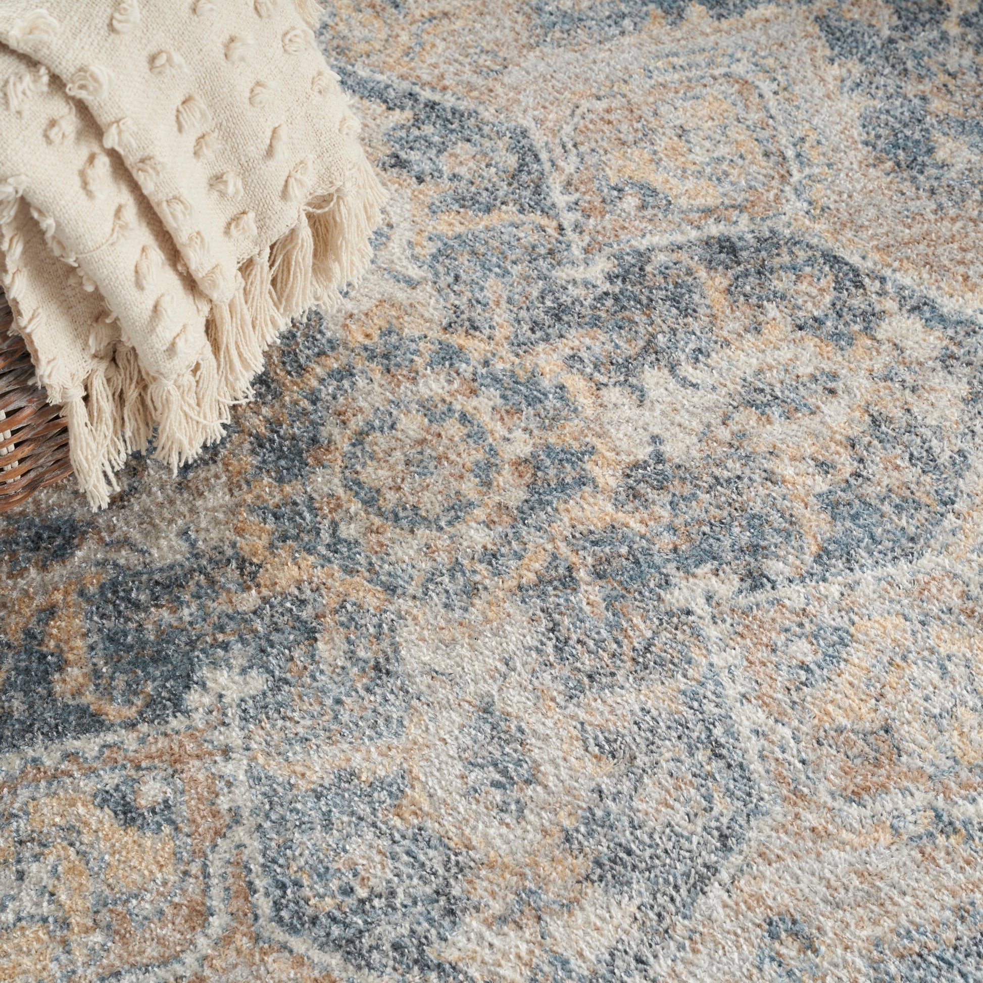 Cream fringed throw draped over a wicker basket on a blue-beige ornate distressed area rug.