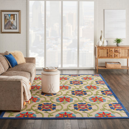 Cozy living room with a vibrant floral rug in blue, red and green; beige sofa, round ottoman and wooden console by windows.