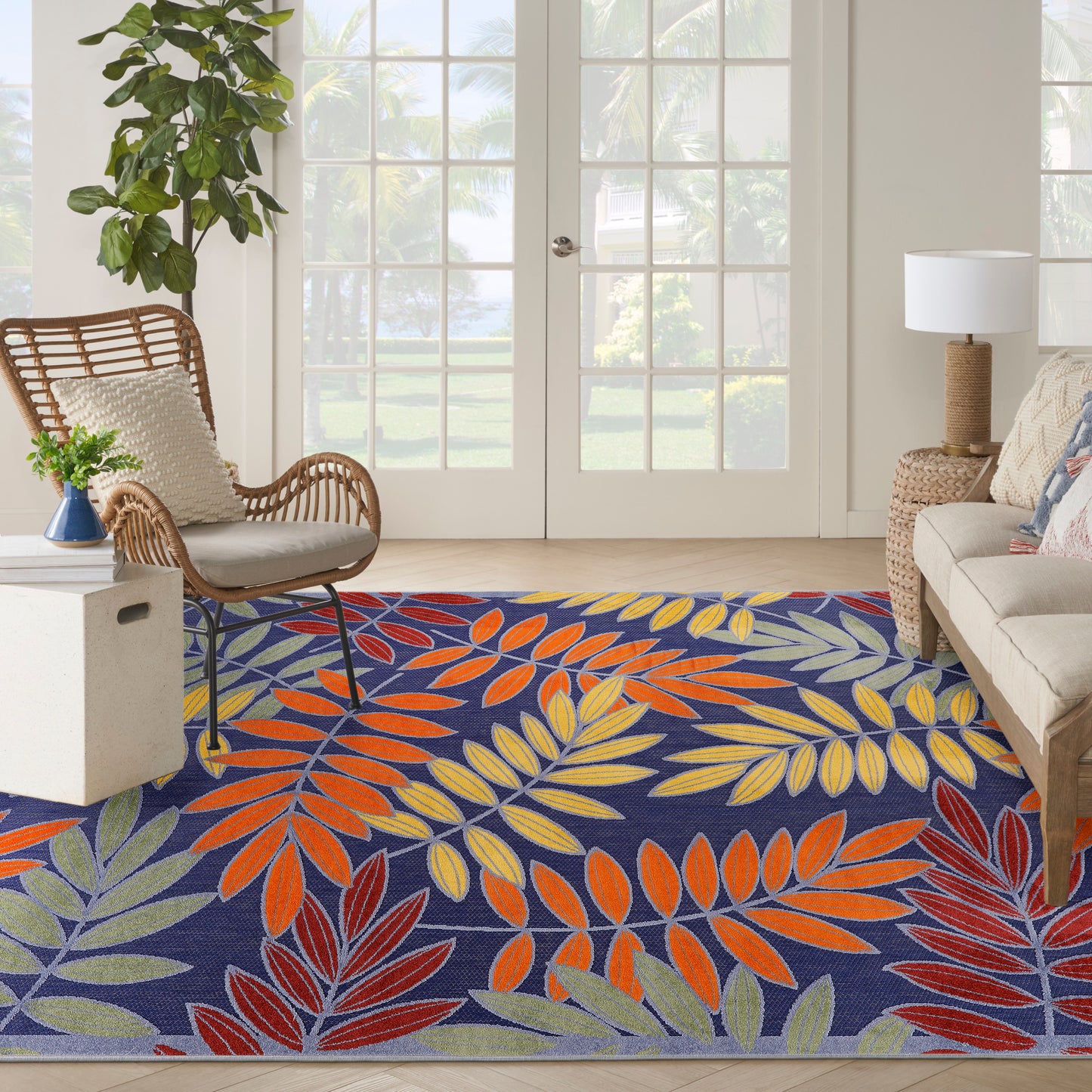 Vibrant woven outdoor rug with multicolored tropical leaf pattern on navy background, in a bright outdoor space with a wicker chair.