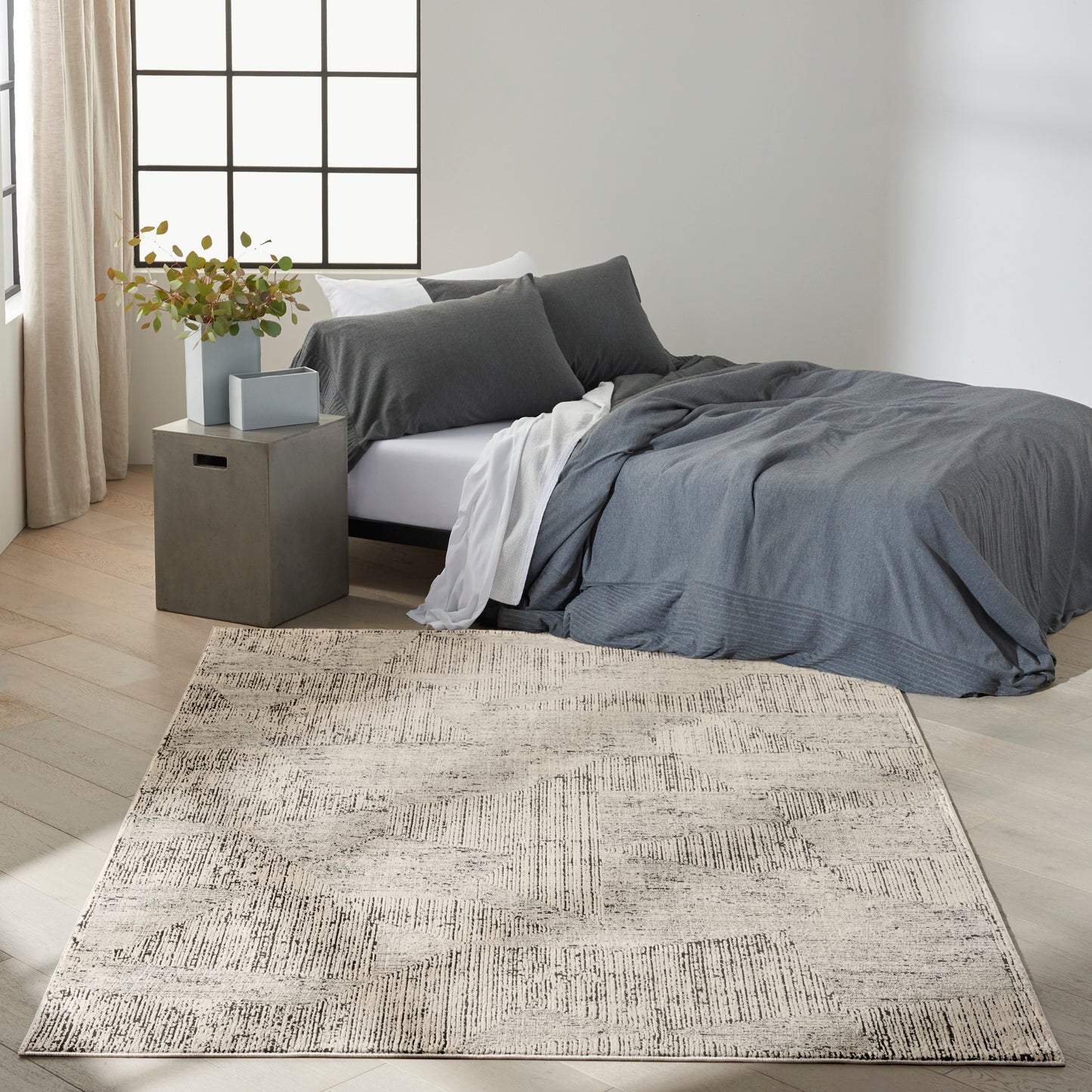 Cozy modern bedroom with gray bedding, beige patterned area rug, square gray nightstand, vase of greenery, large grid window.