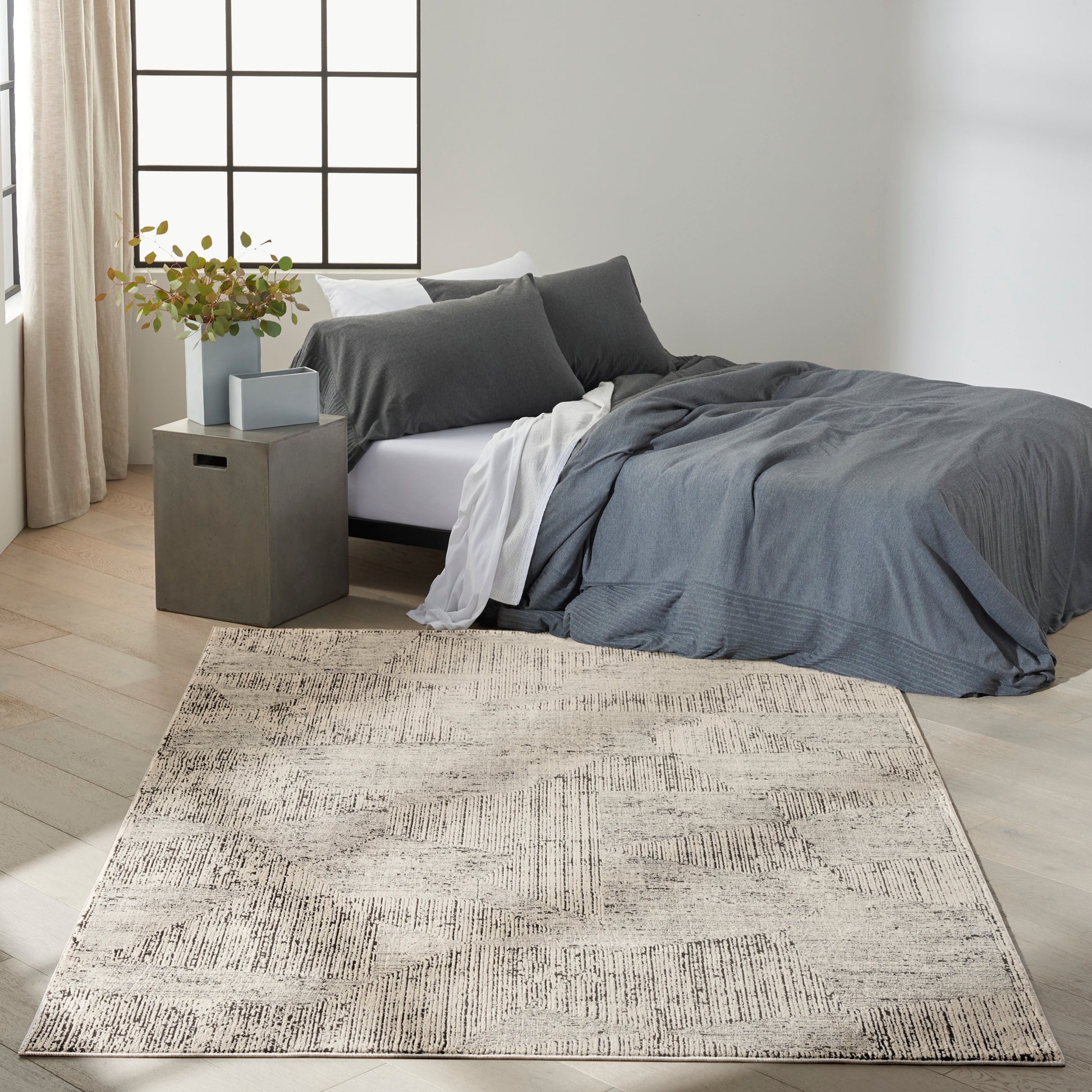 Cozy modern bedroom with gray bedding, beige patterned area rug, square gray nightstand, vase of greenery, large grid window.