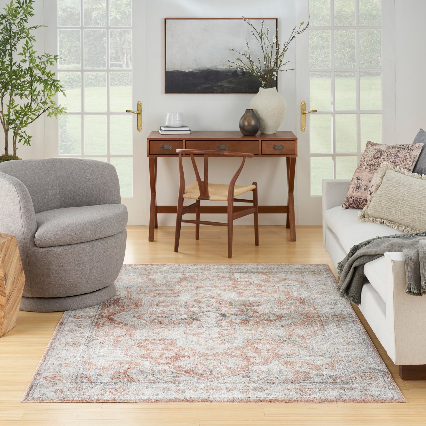 Neutral living room with a vintage patterned rug, oak desk and chair, gray round fabric chair, and beige sofa with pillows.