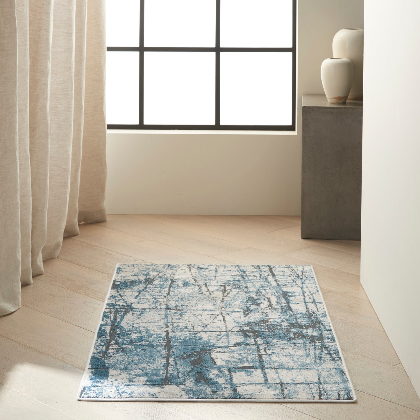Rectangular blue and white abstract-pattern rug on light wood floor in a bright minimalist room with beige curtains.]