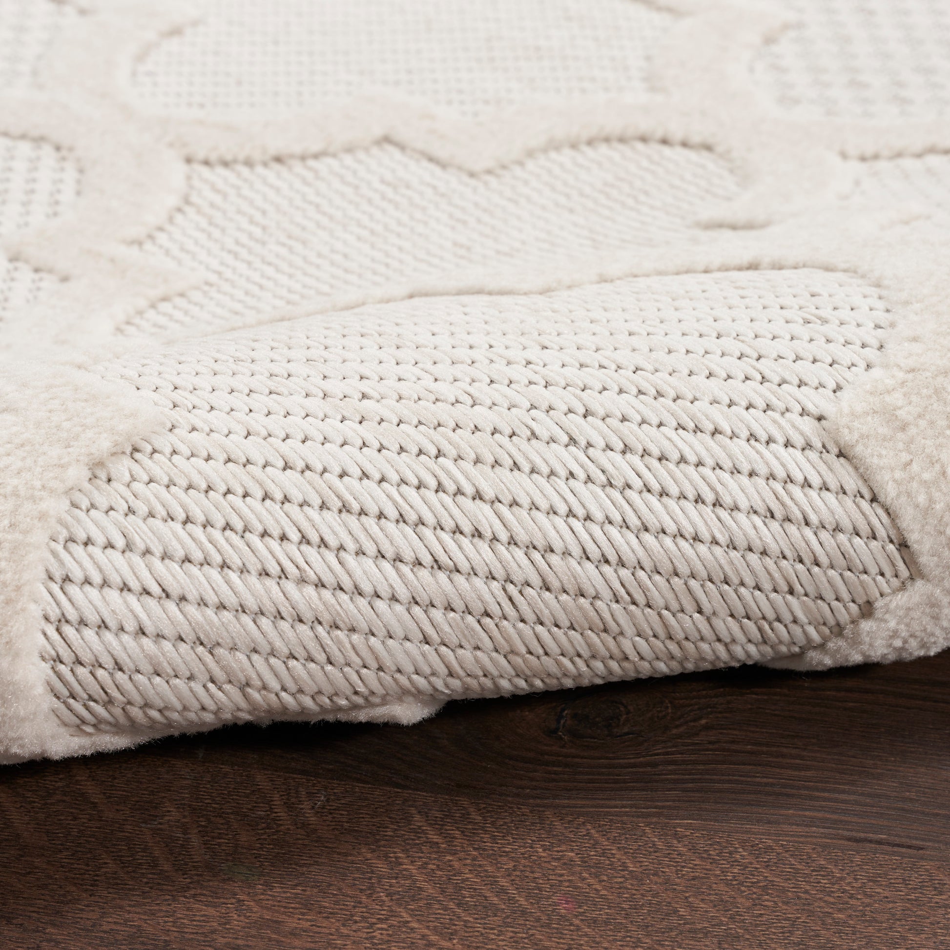 Cream textured rug with a dense twisted pile and raised scroll pattern on a dark wood floor.