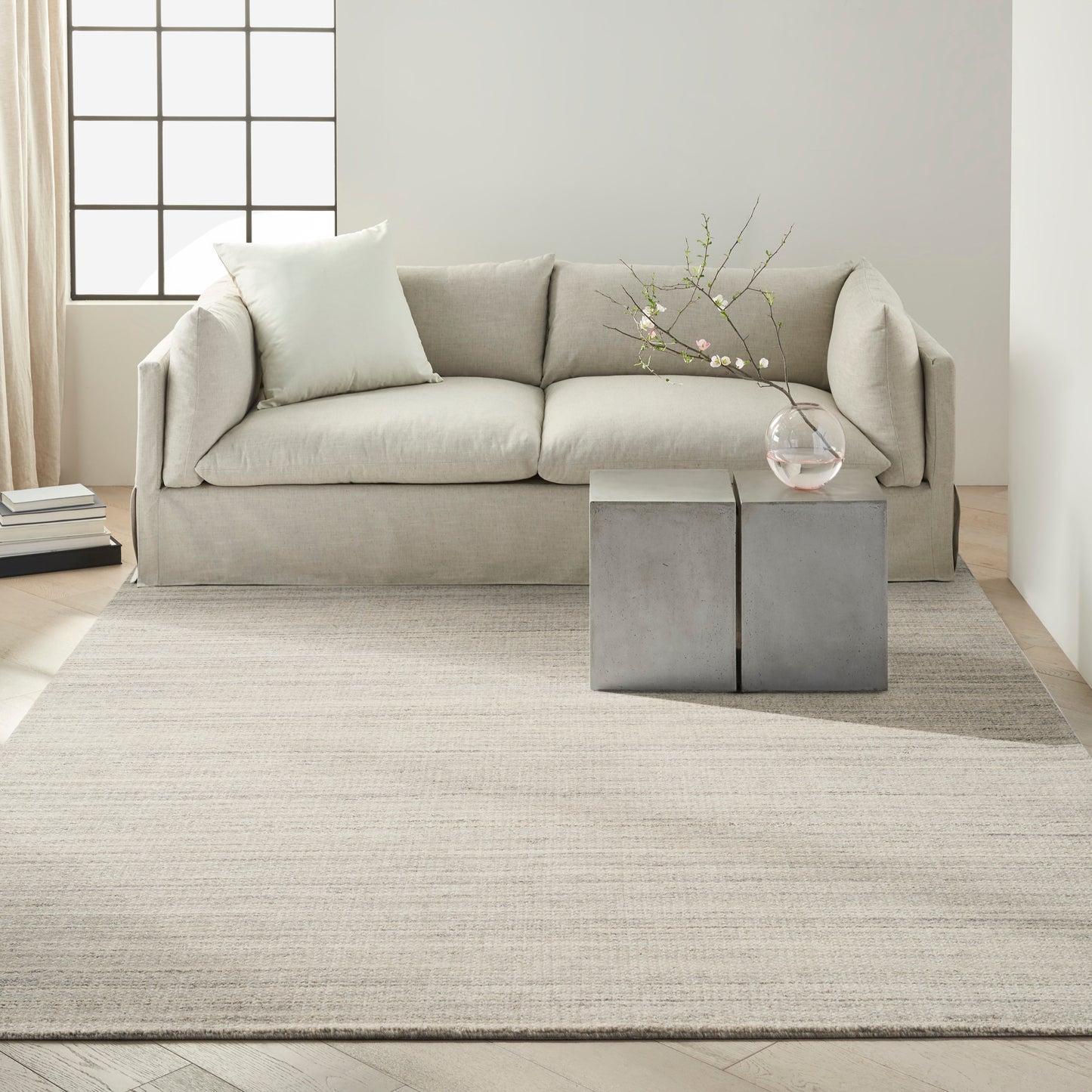 Light gray-beige fabric sofa with plush cushions on a neutral rug; gray cube side tables and a glass vase with branches.