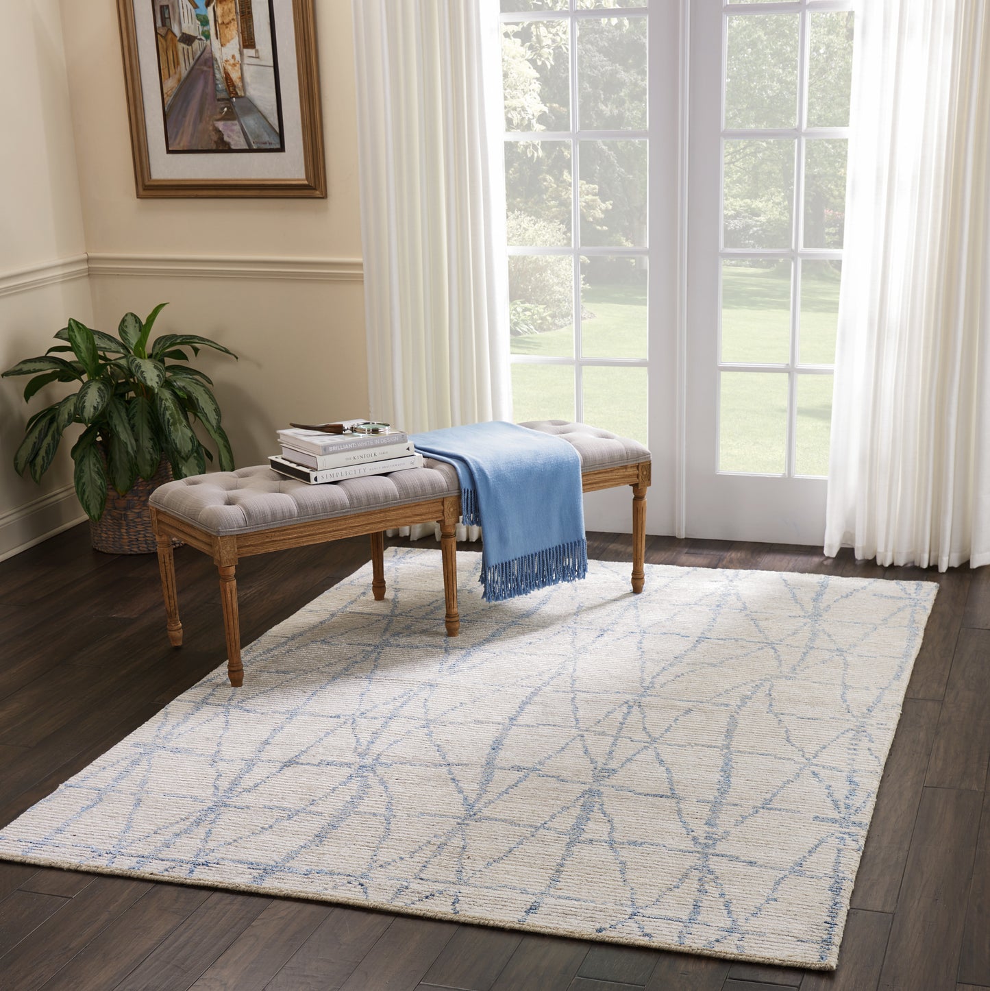 Beige tufted bench with wooden legs, blue throw and books, on a pale geometric rug in a sunlit living room.