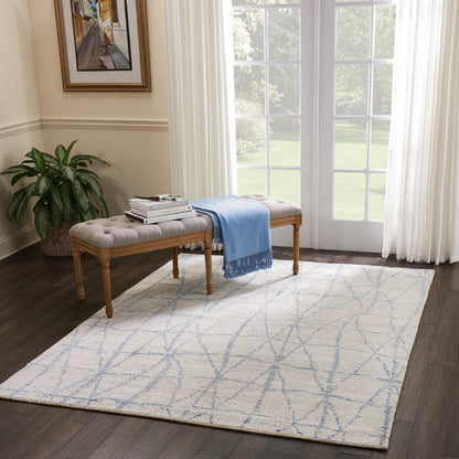 Beige tufted bench with wooden legs, blue throw and books, on a pale geometric rug in a sunlit living room.